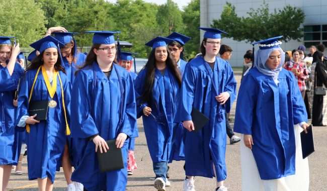 A group of Virtual K-12 graduates in blue caps and gowns walks across a parking lot